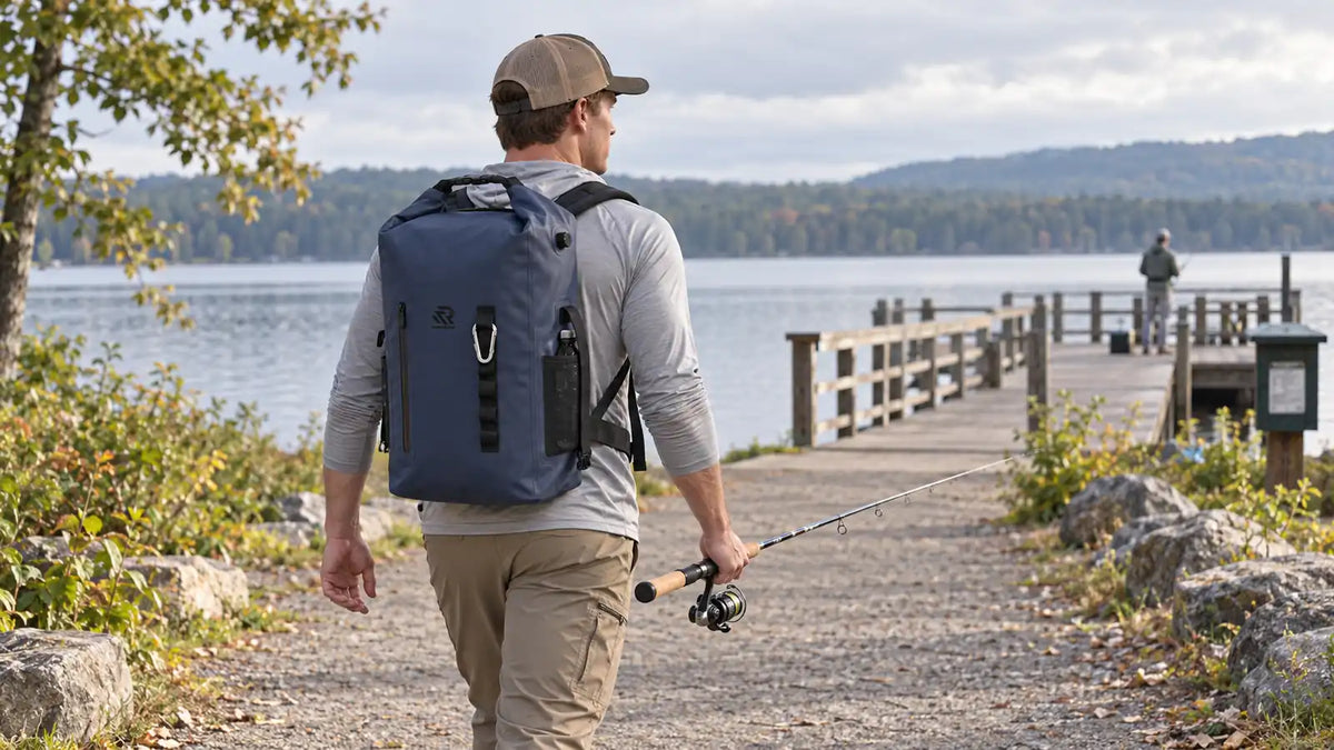An angler wearing a navy RIVONOVA roll-top backpack cooler and carrying a fishing rod toward a lakeside pier